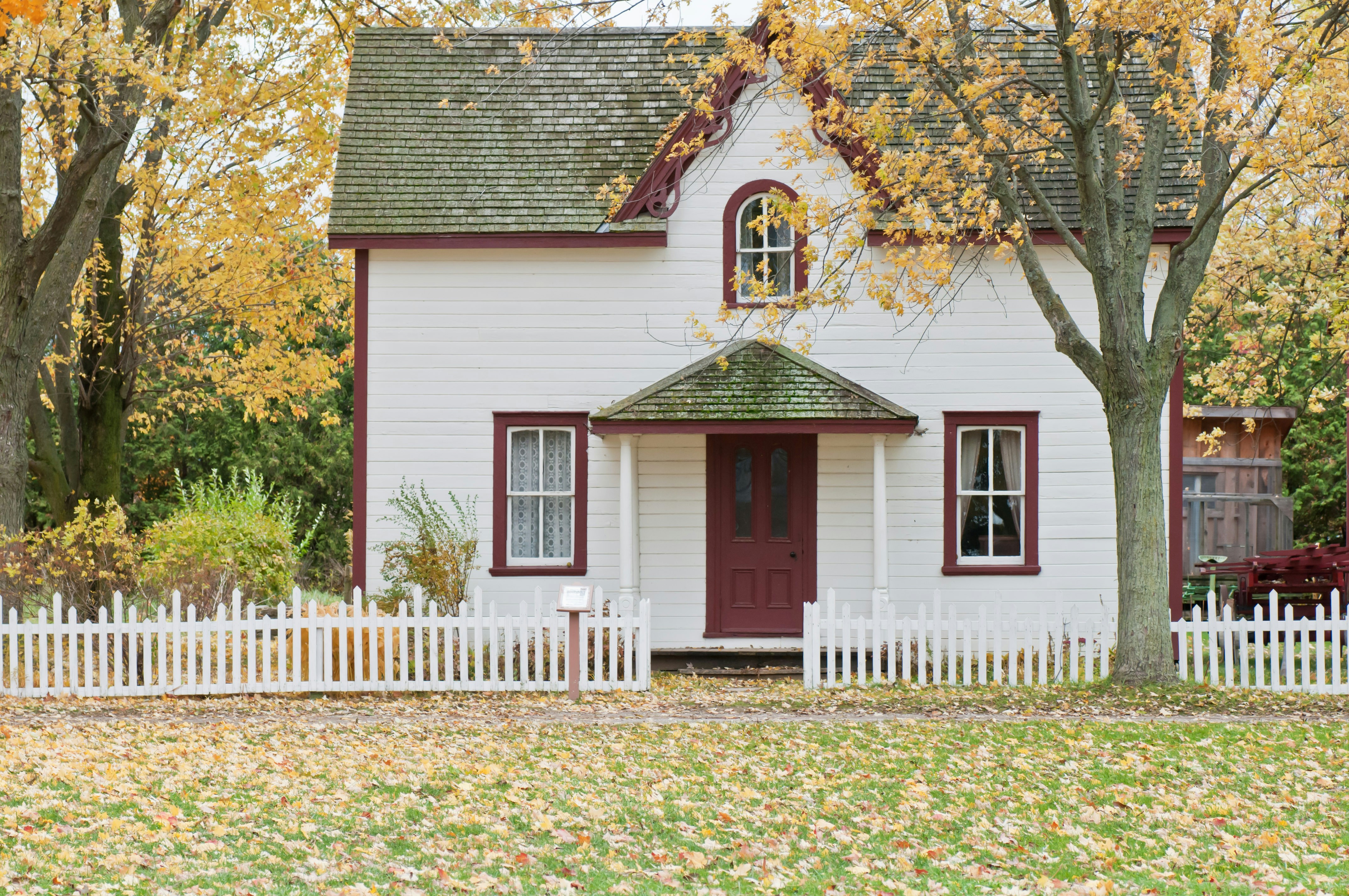 A cozy white house with a red door and a picket fence, surrounded by autumn trees.
